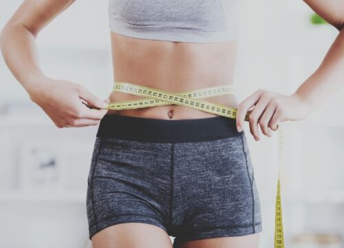 A woman in athletic wear measuring her waist with a yellow tape measure, symbolizing fitness progress and body composition.