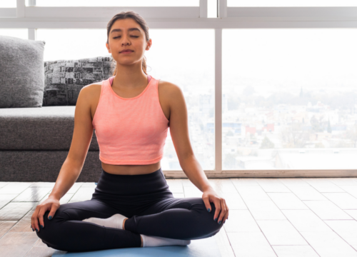 A woman sitting cross-legged in a meditative yoga pose on a mat in a sunlit living room, practicing stress regulation techniques to support her nervous system and body transformation.